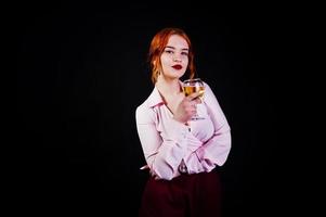 Gorgeous red haired girl in pink blouse and red skirt with glass of wine at hand isolated on black. photo