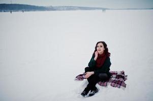 Brunette girl in green sweater and red scarf sitting on plaid outdoor frozen lake on evening winter day. photo