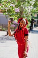 linda y delgada chica afroamericana vestida de rojo con rastas posadas al aire libre en la calle cerca del árbol lila. elegante modelo negro. foto