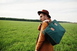 Stylish man in glasses, brown jacket and hat with bag posed on green field. photo