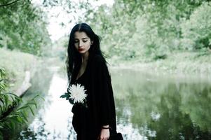 Sensual girl all in black, red lips. Goth dramatic woman hold white chrysanthemum flower at hand against silent lake. photo