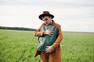 Stylish man in glasses, brown jacket and hat with bag posed on green field. photo