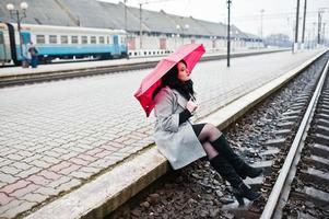 Brunette girl in gray coat with red umbrella in railway station. photo