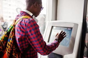 hombre afroamericano en camisa a cuadros con mochila sosteniendo billetera en mano contra atm. viajero hombre negro. foto