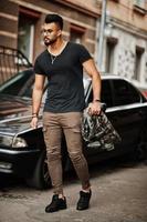 Impresionante y hermoso hombre macho alto de barba árabe con gafas y camiseta negra caminando contra un coche de negocios. foto