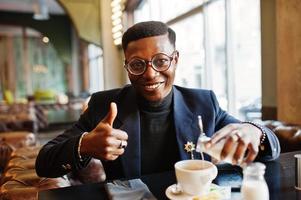 el hombre afroamericano de moda con traje y anteojos vierte crema en el café en la cafetería y muestra el pulgar hacia arriba. foto