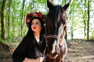 Mystical girl in wreath wear in black with horse in wood. photo