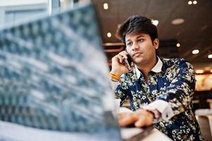 Young male indian freelancer sitting open laptop computer in fast food cafe, handsome asian man speaking phone during work on net-book in comfortable coffee shop. photo