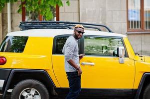 Stylish african american boy on gray sweater and black sunglasses posed on street against yellow car. Fashionable black business man. photo