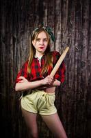 Young funny housewife in checkered shirt and yellow shorts pin up style with kitchen rolling pin on wooden background. photo