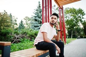 hombre de barba india con estilo en camiseta rosa. modelo indio sentado en un banco de la ciudad y usa su teléfono móvil. foto