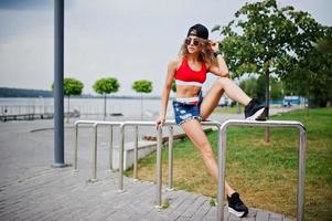 Sexy curly model girl in red top, jeans denim shorts, cup sunglasses and sneakers posed against steel rails. photo