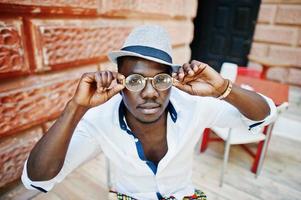 elegante hombre afroamericano con camisa blanca y pantalones de colores con sombrero y gafas al aire libre. chico modelo de moda negro. foto