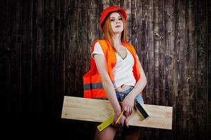 Engineer woman in orange protect helmet and building jacket against wooden background holding board and ruler. photo