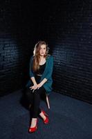 Handsome brunette girl wear on black and green jacket in red high heels, sitting and posing on chair at studio against dark brick wall. Studio model portrait. photo