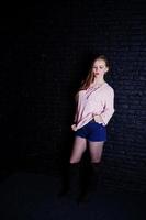 Studio shot of brunette girl at pink sweater with jeans shorts against black brick wall. photo