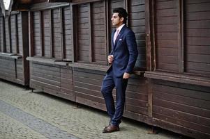 elegante modelo de hombre macho indio con traje y corbata rosa posado contra puestos de madera. foto