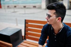 Stylish indian model man in casual clothes, black shirt and sunglasses posed outdoor at street of India and sitting on bench. photo