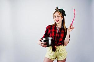 Young funny housewife in checkered shirt and yellow shorts pin up style with saucepan and kitchen spoon isolated on white background. photo