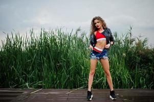 Sexy curly model girl in red top, jeans denim shorts, leather jacket and sneakers posed in the reed. photo