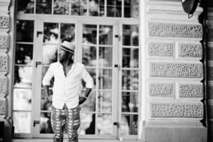 elegante hombre afroamericano con camisa blanca y pantalones de colores con sombrero y gafas al aire libre. chico modelo de moda negro. foto