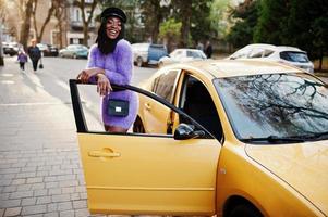 mujer afroamericana con vestido violeta y gorra posada en un auto amarillo. foto