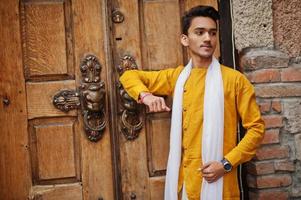 Indian stylish man in yellow traditional clothes with white scarf posed outdoor against old gates. photo