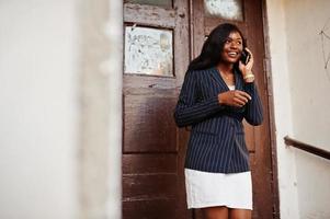 Success stylish african american woman in jacket and skirt against old wooden door with smartphone at hands. photo