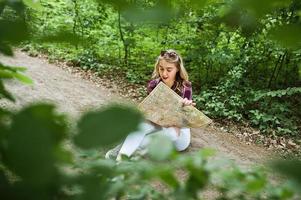 retrato de una joven y hermosa rubia positiva sentada en el suelo con un mapa en las manos en el bosque. foto