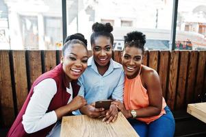Three african american girls sitting on the table of caffe and looking on mobile phone. photo