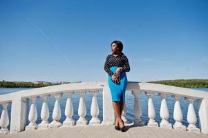 elegante modelo afroamericana con gafas, falda azul y blusa negra posada al aire libre en el puente contra el lago. foto