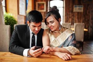 Elegant and fashionable indian friends couple of woman in saree and man in suit posed indoor cafe and looking something on mobile phone. photo
