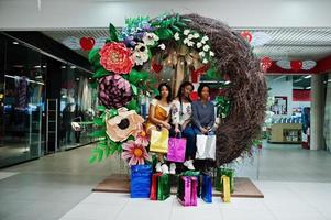 Beautiful three well-dressed afro american girls with colored shopping bags sitting on spring decoration photo zone in mall.