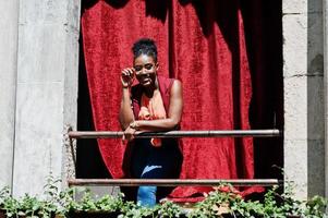 elegante mujer afroamericana posó en balkony con cortina roja al aire libre. foto