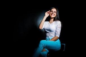 Portrait of an attractive young woman in white top and blue pants sitting posing with her glasses in the dark. photo