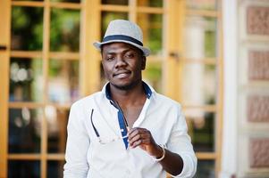 Stylish african american man in white shirt and colored pants with hat and glasses posed outdoor. Black fashionable model boy. photo