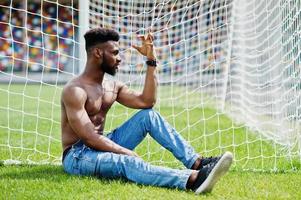 Handsome sexy african american muscular bare torso man at jeans overalls posed at green grass agasinst football gates of stadium field. Fashionable black man portrait. photo