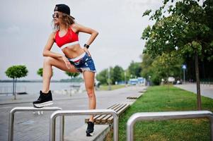 Sexy curly model girl in red top, jeans denim shorts, cup sunglasses and sneakers posed against steel rails. photo