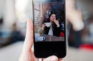 Close up photo of screen mobile phones with brunette girl sitting on cafe with cup of cappuccino, listening music on headphones.