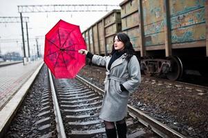 chica morena con abrigo gris con paraguas rojo en la estación de tren. foto