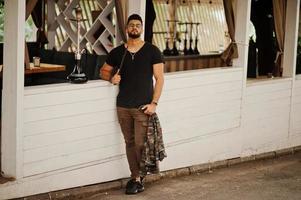 Stylish beard arabian man in glasses and black t-shirt smoking hookah outdoor. Arab model having rest. photo