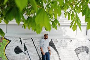 Stylish african american boy on gray sweater and black sunglasses posed at street. Fashionable black guy against graffity wall. photo