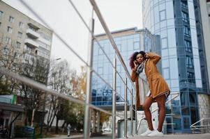 Attractive curly african american woman in brown coat posed near railings against modern multistory building speaking on mobile phone. photo