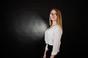 Studio portrait of blonde businesswoman in glasses, white blouse and black skirt against dark background. Successful woman and stylish girl concept. photo