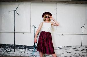 Fashionable and beautiful blonde model girl in stylish red velvet velour skirt, white blouse, hat and sunglasses, posed with phone and earphones. photo
