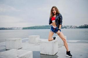 Sexy curly model girl in red top, jeans denim shorts, leather jacket and sneakers posed at stone cubes. photo