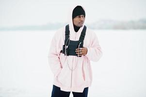 Elegante hombre afroamericano de estilo urbano con capucha rosa posado en un lago congelado en invierno. foto