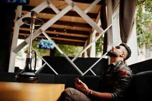 Stylish beard arabian man in glasses and military jacket smoking hookah at street bar. Arab model having rest and looking on phone. photo