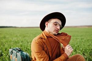 Stylish man in glasses, brown jacket and hat with bag posed on green field. photo