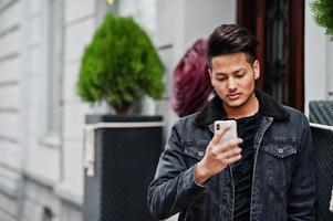 Handsome and fashionable indian man in black jeans jacket posed outdoor and looking at mobile phone. photo